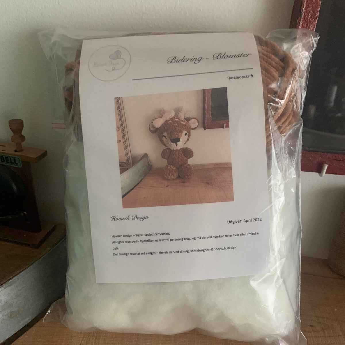 Opskriftsforside for hæklet bamse med lyserøde detaljer, foran pose med garn og fyld